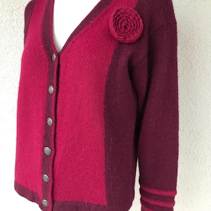 Può includere: Un cardigan di maglia color bordeaux e rosa con una spilla a forma di fiore e chiusura con bottoni.