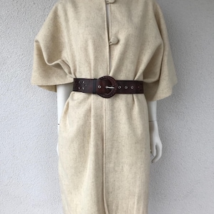 Op de afbeelding: Een crèmekleurige gebreide poncho met een franjezoom en een knoopsluiting. De poncho wordt gedragen met een bruine leren riem met een grote ronde gesp.