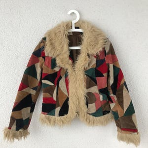 Op de afbeelding: Een vintage patchwork jasje met een kraag en afwerking van imitatiebont. De jas heeft een geometrisch patroon in tinten rood, groen, bruin en zwart. De jas hangt aan een witte hanger tegen een witte achtergrond.