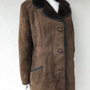 K&ouml;nnte beinhalten: Ein brauner Wildleder-Lammfellmantel mit einem dunkelbraunen Pelzkragen und schwarzen Bes&auml;tzen. Der Mantel hat einen Knopfverschluss und zwei Taschen.