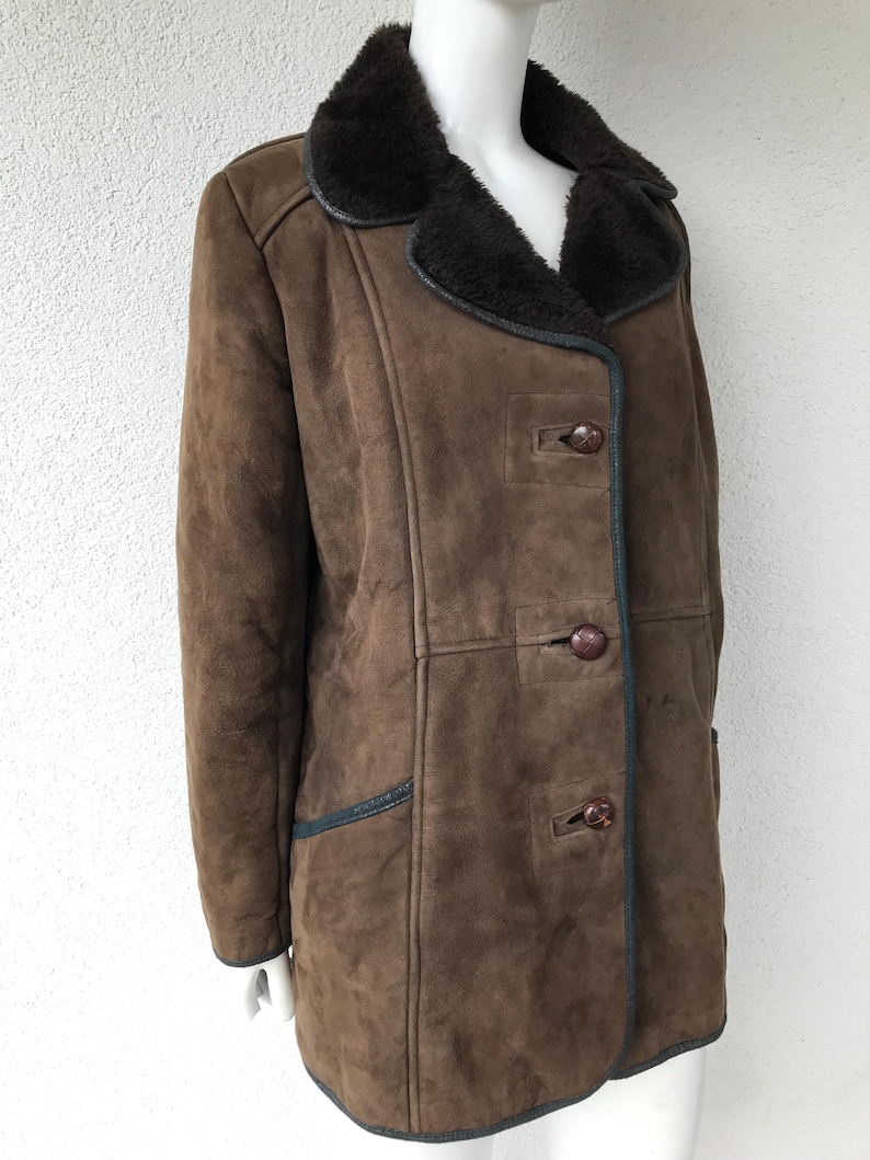 K&ouml;nnte beinhalten: Ein brauner Wildleder-Lammfellmantel mit einem dunkelbraunen Pelzkragen und schwarzen Bes&auml;tzen. Der Mantel hat einen Knopfverschluss und zwei Taschen.