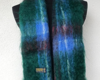 May include: A vibrant, oversized scarf in a plaid pattern, featuring shades of green, blue, and touches of red. The scarf has a soft, fuzzy texture and a small label with text. The scarf is displayed on a black mannequin.