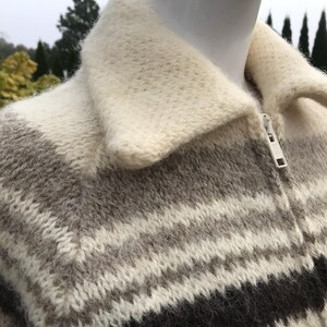 May include: A close-up of a zip-up sweater with a cream-colored collar and a striped pattern in shades of gray and black. The sweater is made of a thick, textured knit fabric.