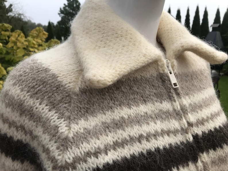 May include: A close-up of a zip-up sweater with a cream-colored collar and a striped pattern in shades of gray and black. The sweater is made of a thick, textured knit fabric.