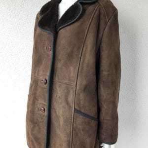 K&ouml;nnte beinhalten: Eine braune Wildleder-Shearling-Jacke mit einem dunkelbraunen Pelzkragen und dunkelbraunen Kn&ouml;pfen. Die Jacke hat einen dunkelbraunen Besatz an den R&auml;ndern und Taschen.