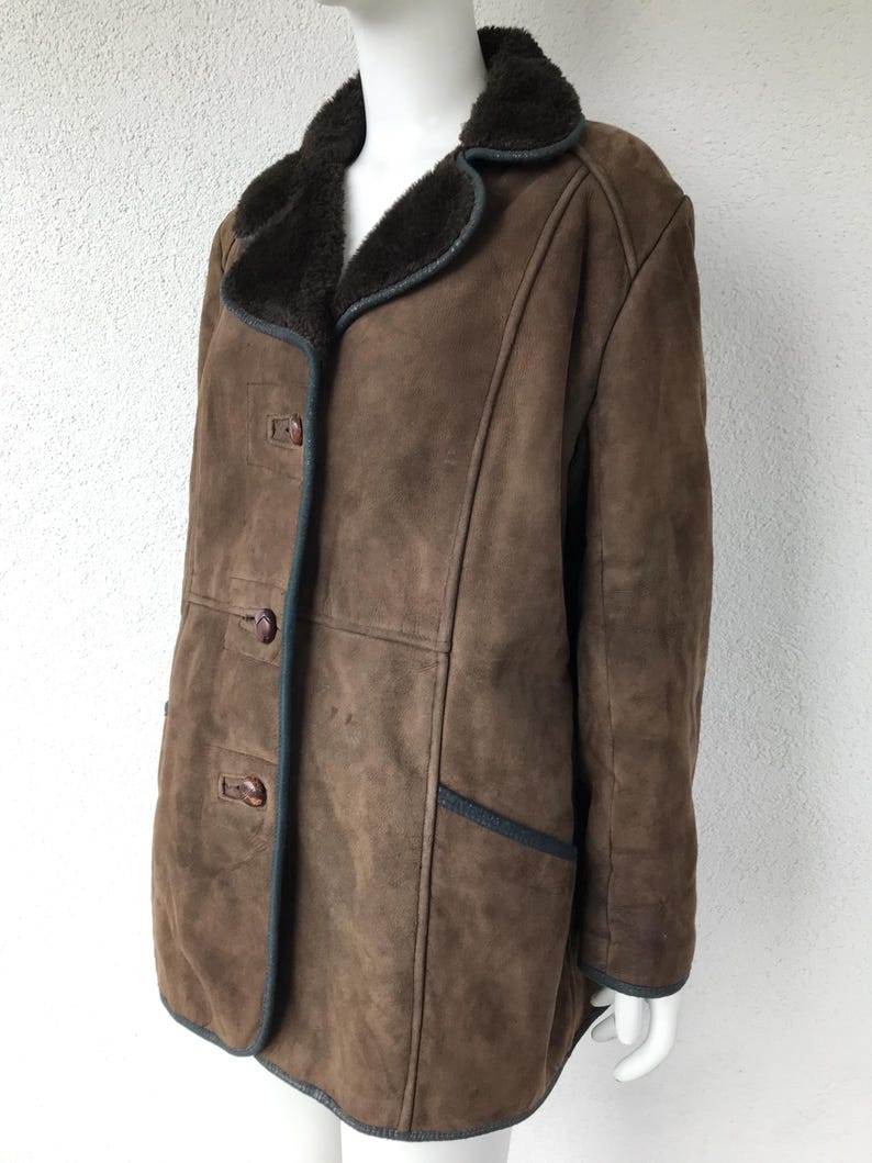 K&ouml;nnte beinhalten: Eine braune Wildleder-Shearling-Jacke mit einem dunkelbraunen Pelzkragen und dunkelbraunen Kn&ouml;pfen. Die Jacke hat einen dunkelbraunen Besatz an den R&auml;ndern und Taschen.