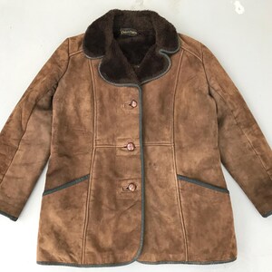 K&ouml;nnte beinhalten: Eine braune Wildlederjacke mit einem braunen Kunstfellkragen und drei Kn&ouml;pfen. Die Jacke hat einen dunkelgr&uuml;nen Besatz und zwei Taschen.