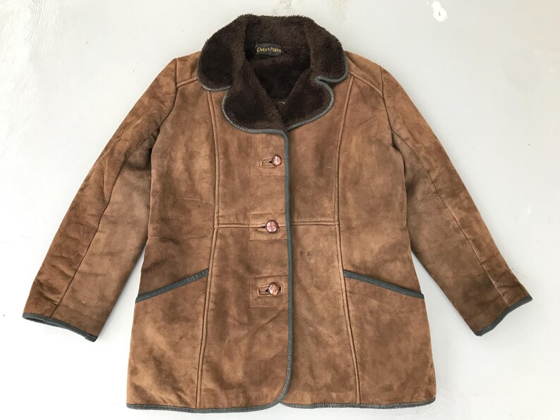 K&ouml;nnte beinhalten: Eine braune Wildlederjacke mit einem braunen Kunstfellkragen und drei Kn&ouml;pfen. Die Jacke hat einen dunkelgr&uuml;nen Besatz und zwei Taschen.