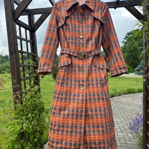 Op de afbeelding: Een lange trenchcoat met een levendig oranje en donkergrijs ruitpatroon. De jas is voorzien van een puntige kraag, lange mouwen, een knoopsluiting aan de voorzijde en een bijpassende ceintuur met een grote, ronde gesp in de taille.