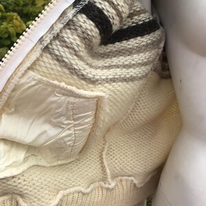 May include: Cream-colored knitted jacket with a white zipper and a striped collar in shades of brown and beige. The jacket features a white satin-like lining and a pocket. The bottom hem has a scalloped edge.
