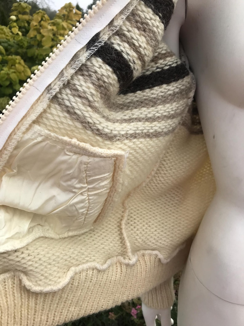 May include: Cream-colored knitted jacket with a white zipper and a striped collar in shades of brown and beige. The jacket features a white satin-like lining and a pocket. The bottom hem has a scalloped edge.