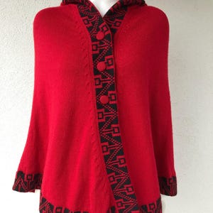 Op de afbeelding: Rode gebreide poncho met capuchon en zwarte geometrische rand. De poncho heeft een knoopsluiting en een capuchon.