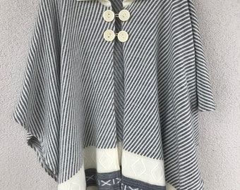 May include: A white and grey striped knitted poncho with a white collar and four white buttons. The poncho has a white embroidered design on the bottom hem.