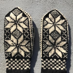 May include: A pair of black and white knitted mittens. Each mitten features a large, symmetrical snowflake design in white against a black background. The cuffs have a geometric pattern. The mittens are likely made of wool.