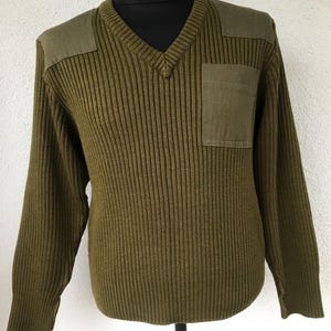 May include: Olive green V-neck sweater with a ribbed knit design. It has reinforced shoulder patches and a single square pocket on the chest. The long-sleeved sweater is displayed on a black mannequin.