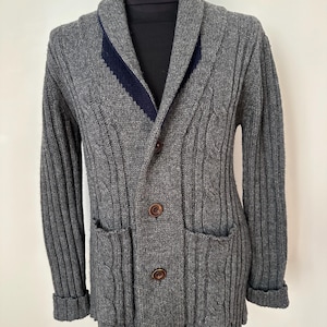 Puede incluir: Un cárdigan gris de punto trenzado con cuello chal y dos bolsillos delanteros. El suéter tiene un acento azul marino en el cuello y botones marrones. Las mangas están dobladas en las muñecas.