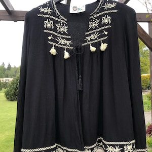 Peut inclure: Haut noir à manches longues avec broderies florales crème sur l'encolure, les poignets et l'ourlet. Le haut a un col en V avec une attache à pompon et de petits pompons. Le vêtement est suspendu à un cintre noir.