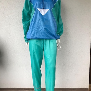 Può includere: Tuta vintage in nylon blu e verde acqua. La giacca ha una mezza zip, dettagli bianchi e la scritta "new-line". I pantaloni hanno strisce riflettenti. L'outfit è esposto su un manichino.