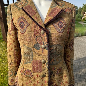 Puede incluir: Una chaqueta de manga larga con un patrón de tapiz en tonos oro, mostaza y multicolor con motivos florales y geométricos. La chaqueta tiene un escote en V, un ribete trenzado de color morado oscuro en la abertura frontal y cuatro botones redondos de latón.