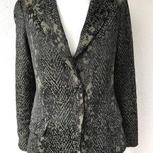 Pode incluir: Um blazer cinza escuro com um padrão de pele de cobra. O blazer tem lapelas entalhadas, dois bolsos frontais e um fecho de um botão. O tecido tem um brilho subtil, com tons de preto e verde-oliva.