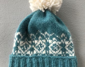 May include: A teal and white knit hat with a large white pom-pom on top. The hat features a ribbed brim and a geometric pattern in white. The hat is made of a soft, textured material, perfect for cold weather.