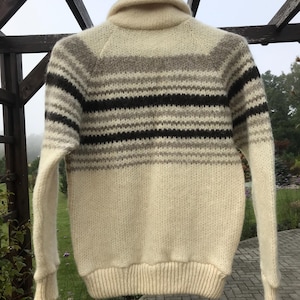 May include: Cream-colored knit sweater with a shawl collar and horizontal stripes in shades of brown, gray, and black. The sweater has long sleeves and a ribbed hem and cuffs. The garment is hanging outdoors.