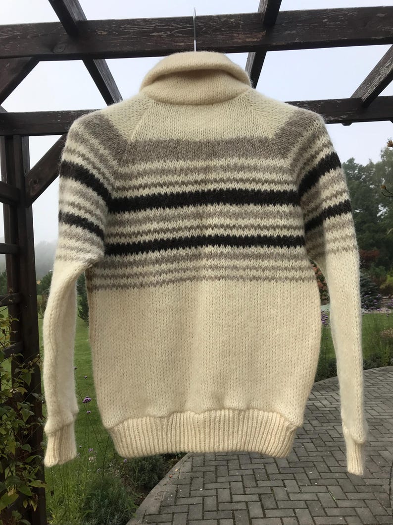 May include: Cream-colored knit sweater with a shawl collar and horizontal stripes in shades of brown, gray, and black. The sweater has long sleeves and a ribbed hem and cuffs. The garment is hanging outdoors.