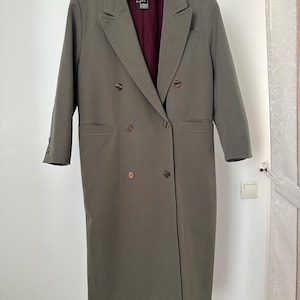 May include: A long, olive green double-breasted coat with a classic design. The coat features a notched lapel, multiple brown buttons, and a burgundy lining. It is hanging on a wooden hanger against a white wall.