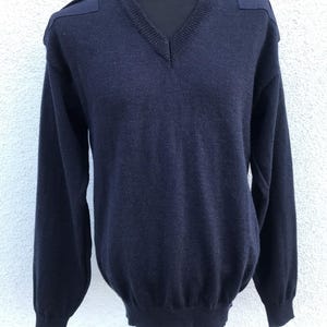 May include: Navy blue V-neck jumper with shoulder epaulettes. The long-sleeved jumper is displayed on a black mannequin. The jumper has a ribbed hem and cuffs. The fabric appears to be a medium-weight knit.