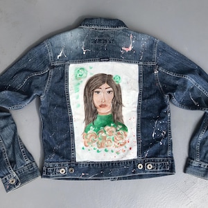Peut inclure: Veste en jean avec un portrait peint à la main au dos. L'œuvre représente une femme aux cheveux bruns et un haut vert, entourée d'éléments floraux. La veste présente des éclaboussures de peinture et des fermetures à boutons.