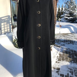 Puede incluir: Un abrigo largo negro con cuello de piel sintética y botones dorados. El abrigo tiene mangas largas y un diseño clásico. El abrigo se exhibe en un maniquí en un entorno nevado al aire libre.