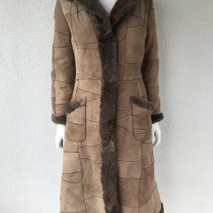 May include: A long, brown shearling coat with a patchwork design. The coat features a large collar, pockets, and dark brown trim. The coat is displayed on a mannequin.