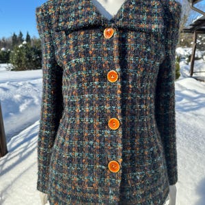 Könnte beinhalten: Vintage Tweed-Jacke in einem dunkelblauen, teal- und orangefarbenen Karomuster. Die Jacke hat einen klassischen Kragen, lange Ärmel und drei große orangefarbene Knöpfe. Die Jacke wird auf einer Schaufensterpuppe in einer verschneiten Umgebung präsentiert.