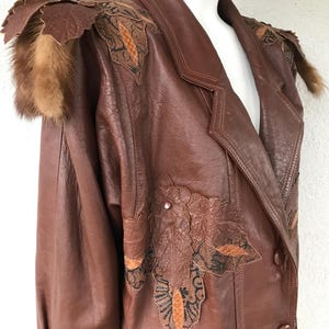 Op de afbeelding: Een bruin leren jack met ingewikkelde bloemmotieven en bontbeslag op de schouders.