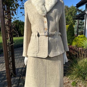 Op de afbeelding: Een beige tweedelig pak bestaande uit een jasje met lange mouwen en een bijpassende rok. Het jasje heeft een witte kraag van imitatiebont, drie knopen aan de voorzijde, twee voorzakken en een smalle riem met een zilverkleurige gesp in de taille.