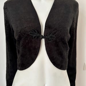 Puede incluir: Chaqueta bolero negra con mangas largas y cuello en V. La chaqueta presenta un cierre único con dos nudos decorativos. La tela tiene un sutil patrón texturizado, agregando interés visual a la prenda. La chaqueta se muestra en un maniquí.
