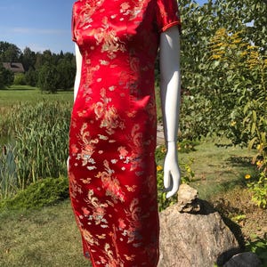 Puede incluir: Vestido rojo de manga corta con cuello mandarín y estampado de dragones. El vestido es de tejido brillante y llega a la rodilla. El vestido se exhibe en un maniquí en un entorno al aire libre.