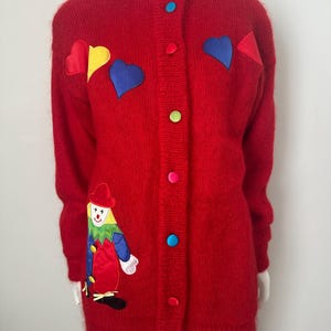 Peut inclure: Un cardigan rouge vif avec un col montant et des boutons colorés. Le pull présente des cœurs appliqués en jaune, bleu et rouge, ainsi qu'un appliqué de clown. Le pull à manches longues est présenté sur un mannequin.