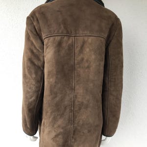 K&ouml;nnte beinhalten: Eine braune Wildlederjacke mit einem schwarzen Kunstfellkragen. Die Jacke hat einen langen, geraden Schnitt und einen Knopfverschluss.