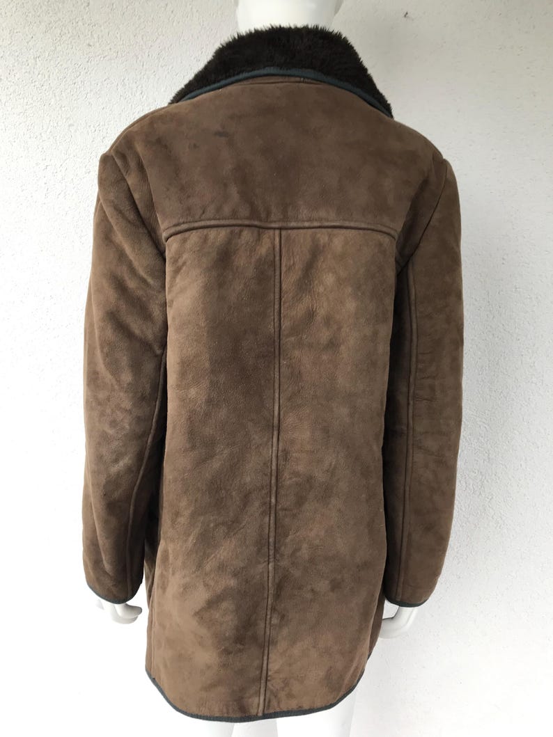 K&ouml;nnte beinhalten: Eine braune Wildlederjacke mit einem schwarzen Kunstfellkragen. Die Jacke hat einen langen, geraden Schnitt und einen Knopfverschluss.