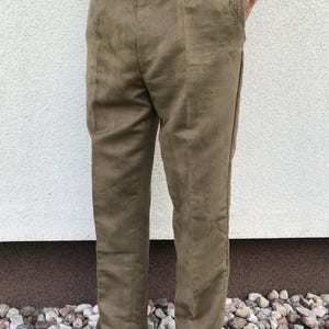 Könnte beinhalten: Hellbraune Hose mit geradem Bein. Die Hose hat einen Knopfverschluss und Gürtelschlaufen. Ein kleines gesticktes Design ist in der Nähe der Tasche zu sehen. Das Material ist weich und Wildleder-ähnlich.
