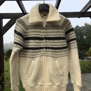 May include: A vintage zip-up sweater with a cream-colored body and a striped pattern in shades of brown and gray. The sweater features a wide collar, two front pockets, and ribbed cuffs and hem. The zipper is silver.