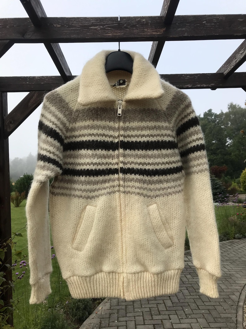 May include: A vintage zip-up sweater with a cream-colored body and a striped pattern in shades of brown and gray. The sweater features a wide collar, two front pockets, and ribbed cuffs and hem. The zipper is silver.