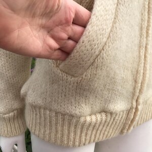 May include: Cream-colored knitted zip-up sweater with a ribbed hem and cuffs. The sweater features a front pocket and a textured knit design. The zipper is visible, and the overall style is casual and warm.