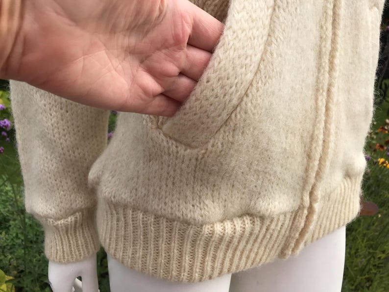 May include: Cream-colored knitted zip-up sweater with a ribbed hem and cuffs. The sweater features a front pocket and a textured knit design. The zipper is visible, and the overall style is casual and warm.