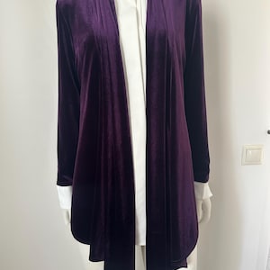 May include: A deep purple velvet cardigan with long sleeves and a draped front. The cardigan is layered over a white collared shirt with white cuffs. The garment is displayed on a mannequin.