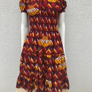 May include: A maroon off-the-shoulder dress with a yellow and white geometric print. The dress has a smocked bodice and a tiered skirt with a lace trim.