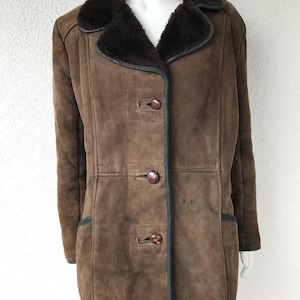 K&ouml;nnte beinhalten: Ein brauner Wildleder-Shearling-Mantel mit einem dunkelbraunen Pelzkragen und vier braunen Kn&ouml;pfen. Der Mantel hat einen dunkelbraunen Besatz an Kragen, Taschen und Saum.