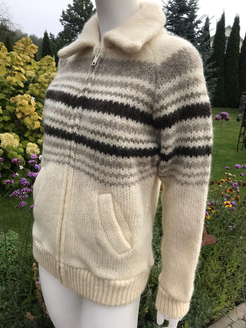 May include: A cream-colored zip-up sweater with horizontal stripes in shades of brown and gray. The sweater has a collared neckline, two front pockets, and ribbed cuffs and hem. The zipper is silver.