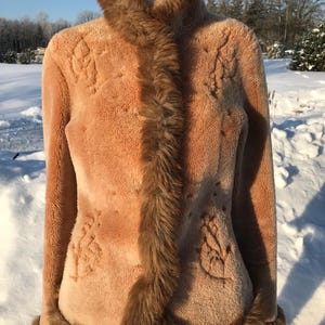 Puede incluir: Un abrigo de piel sintética color canela con cuello alto y ribete marrón. El abrigo presenta diseños florales en relieve y se exhibe en un maniquí. El fondo muestra un paisaje nevado con árboles.